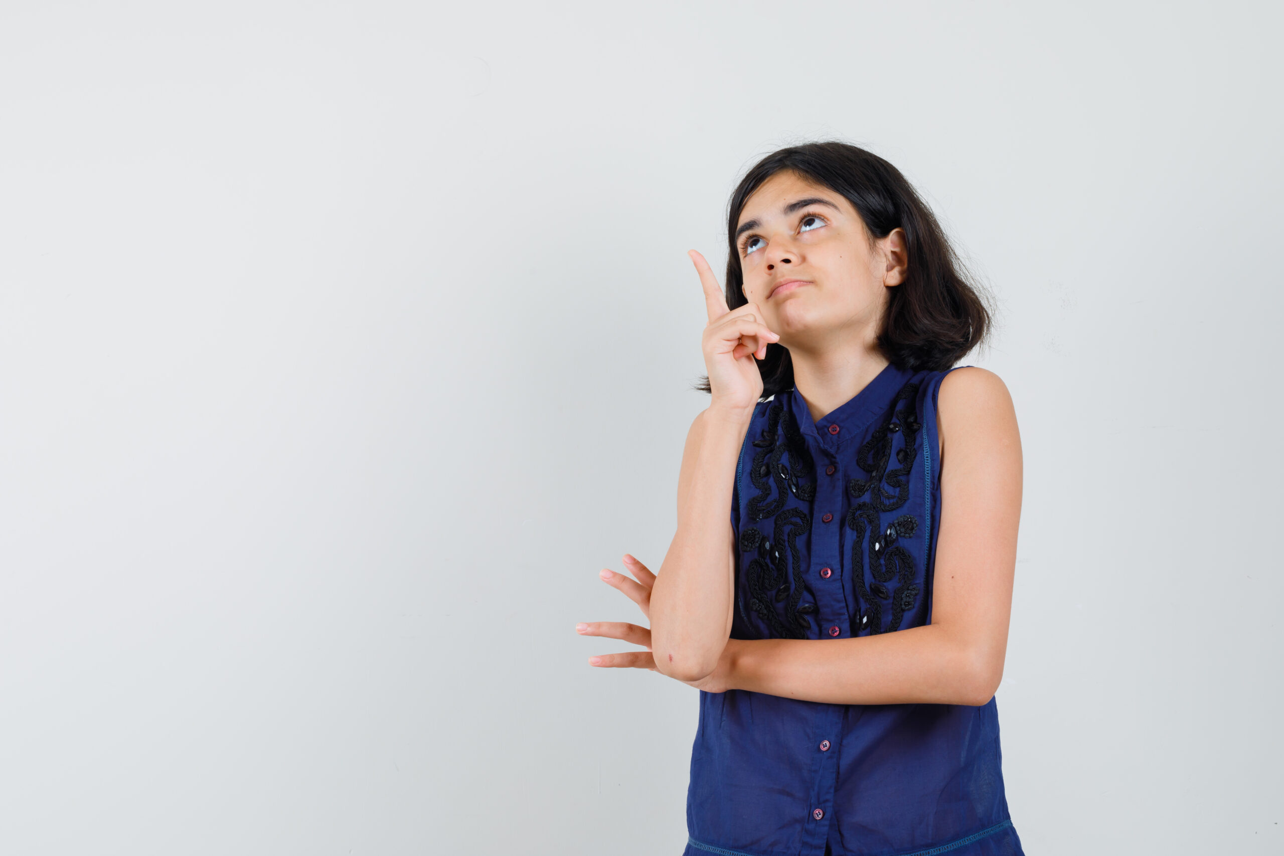 Little Girl Pointing Up In Blue Blouse And Looking Curious. Front View.